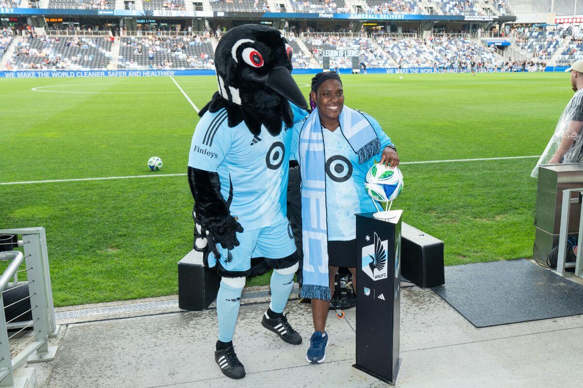 Woman in MNUFC team jersey with loon mascot standing in front of soccer field