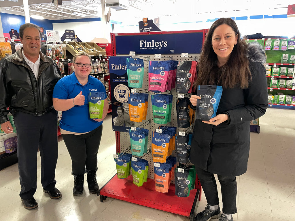 Finley's team stands next to an endcap at PetCo with Finley's Biscuits, Training Bites, and Benefit Bars featured on the endcap. A Finley's Ambassador is standing wearing a blue shirt smiling and holding a thumb up with a bag of treats in her hand. 