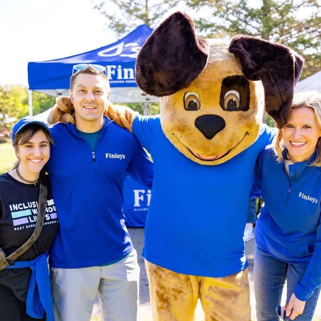 People in Finley's apparel standing with a dog mascot at an event with a blue tent in the background