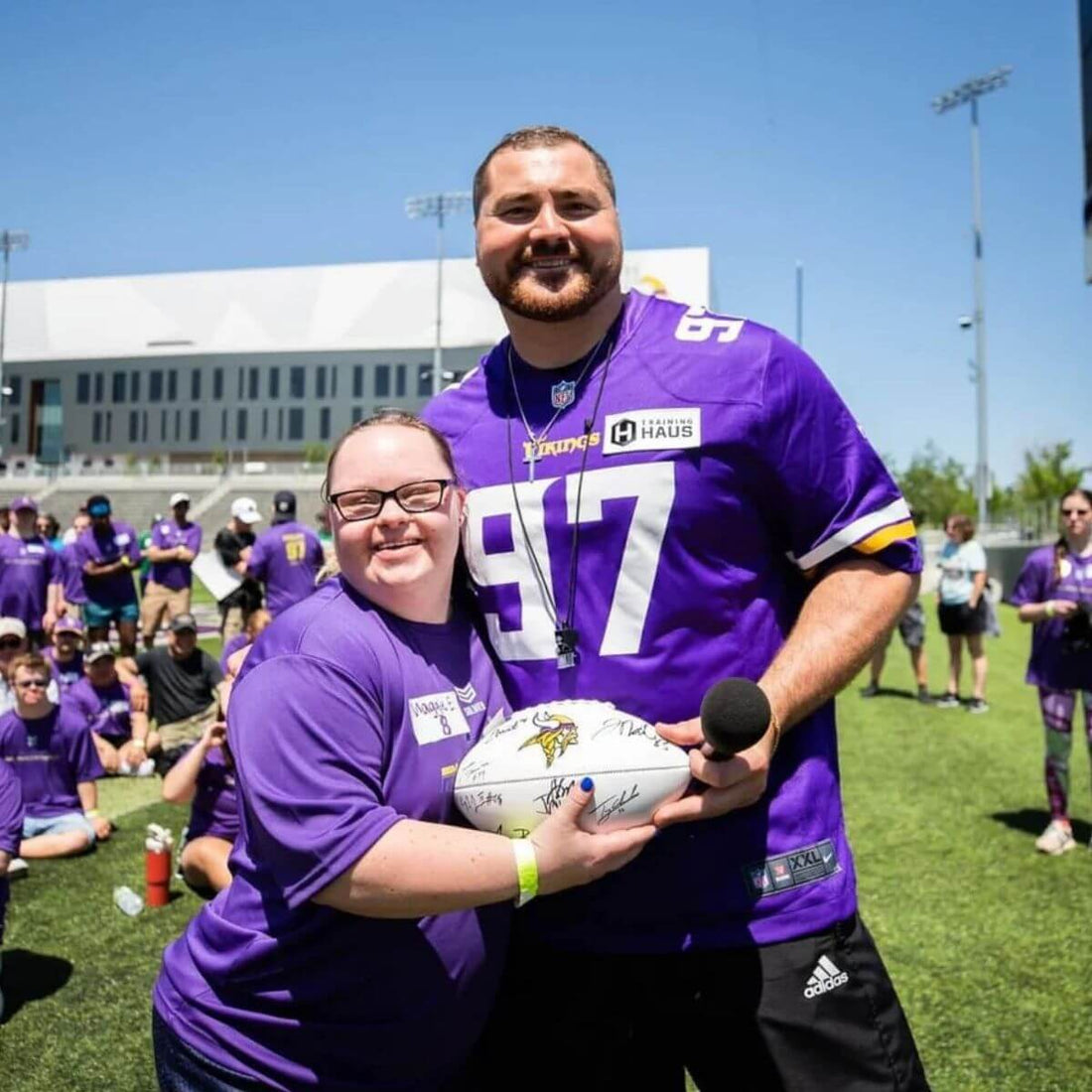 Two people in purple sports jerseys holding a signed Vikings football on a sports field
