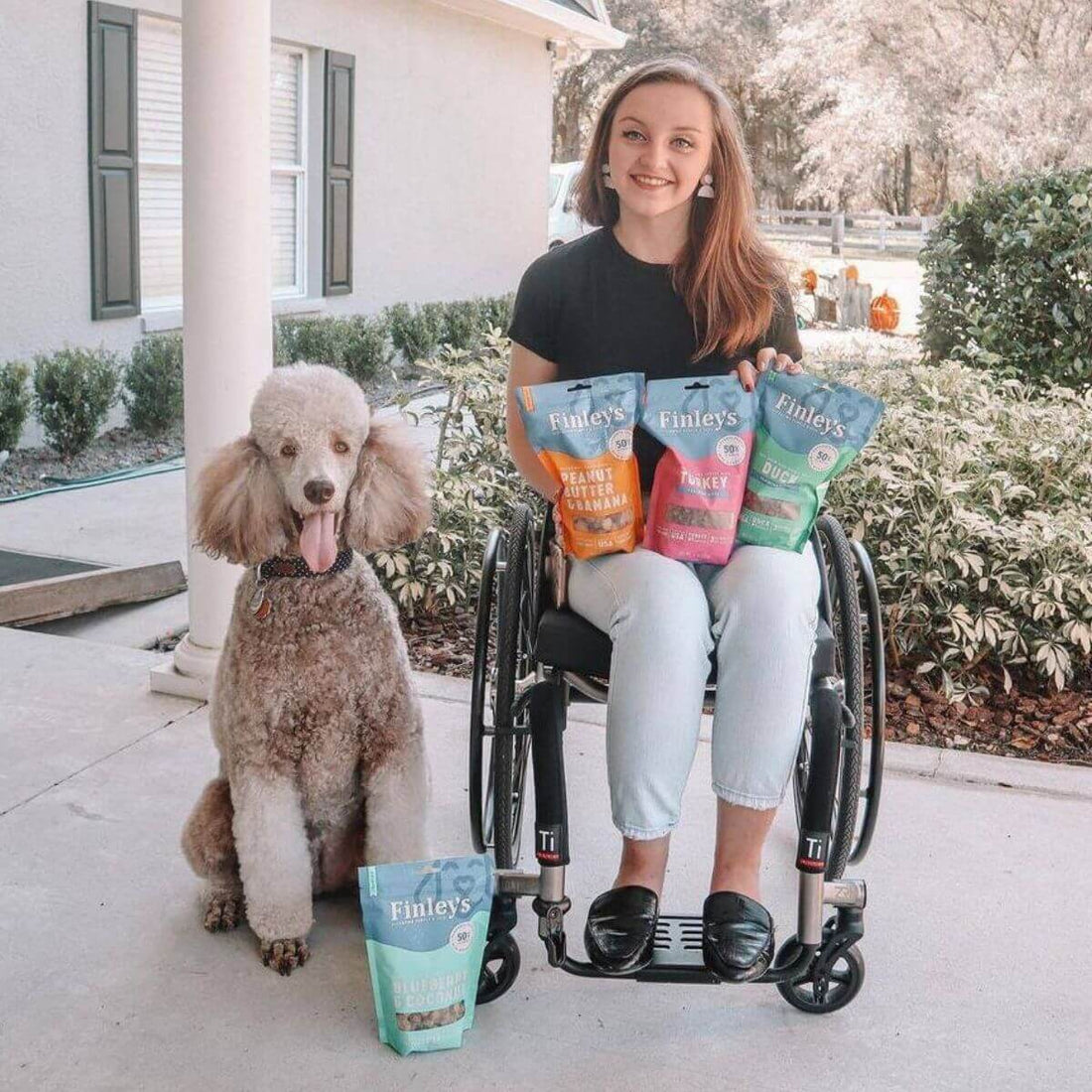 Person in a wheelchair holding bags of Finley's treats with a dog sitting next to them